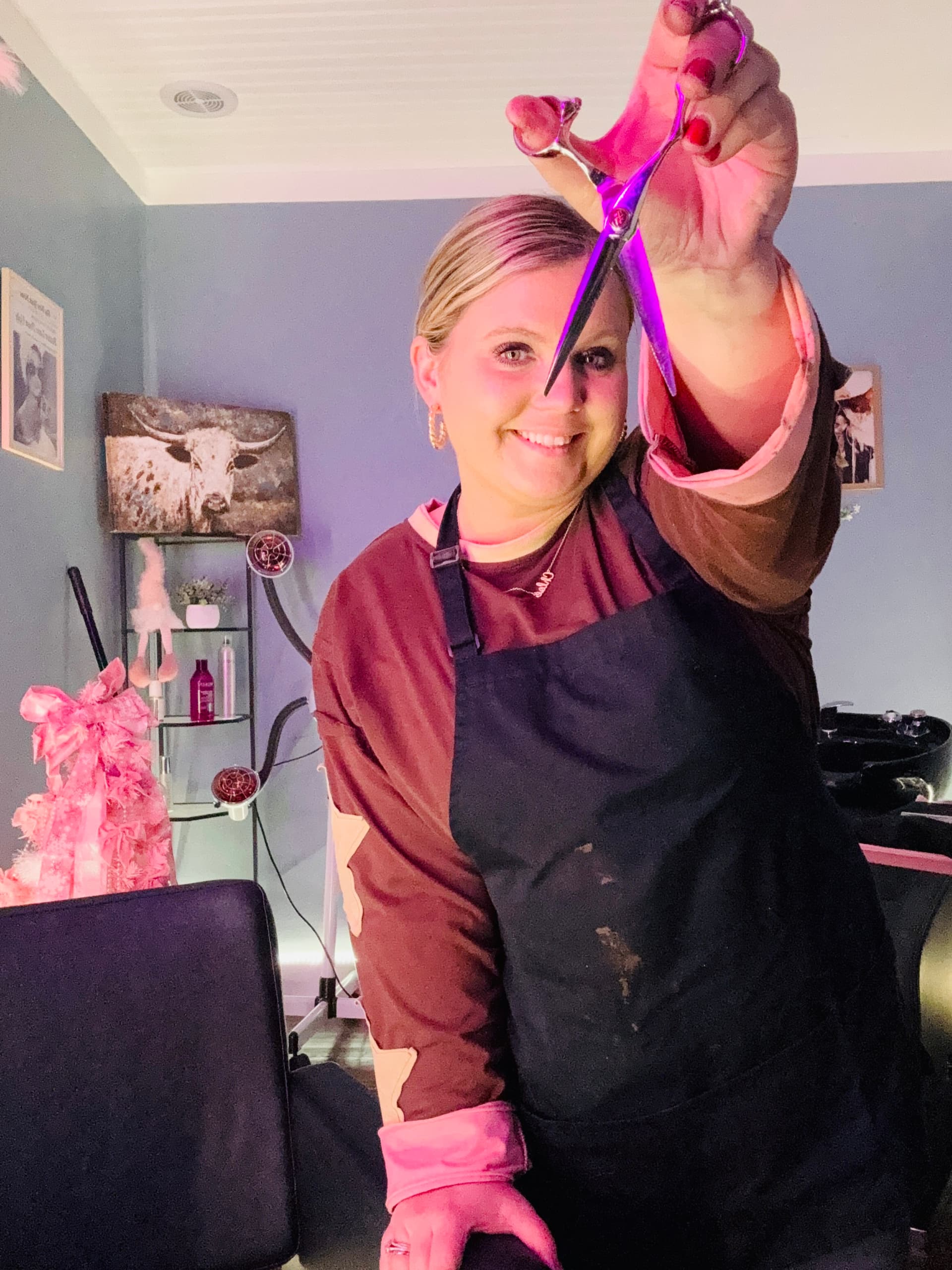 Brooke Holland, solo hair stylist, smiling in her salon holding her shears, wearing her stylist apron.